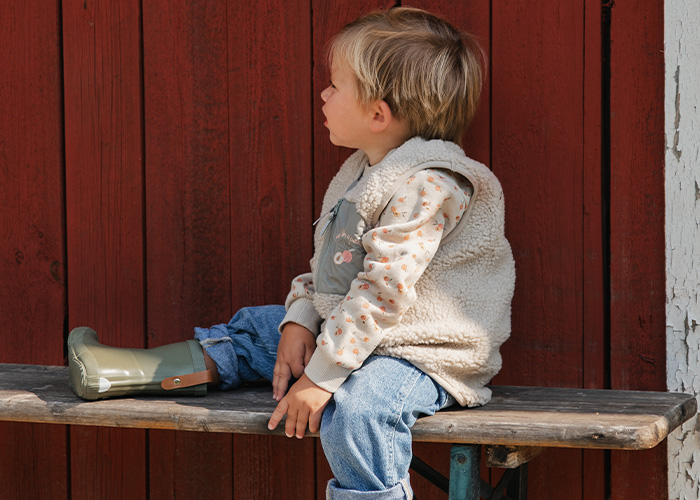 Lapsi istuu penkillä, pukeutuneena liiviin ja kumisaappaisiin, tekstillä 'Kenza X Viking'.