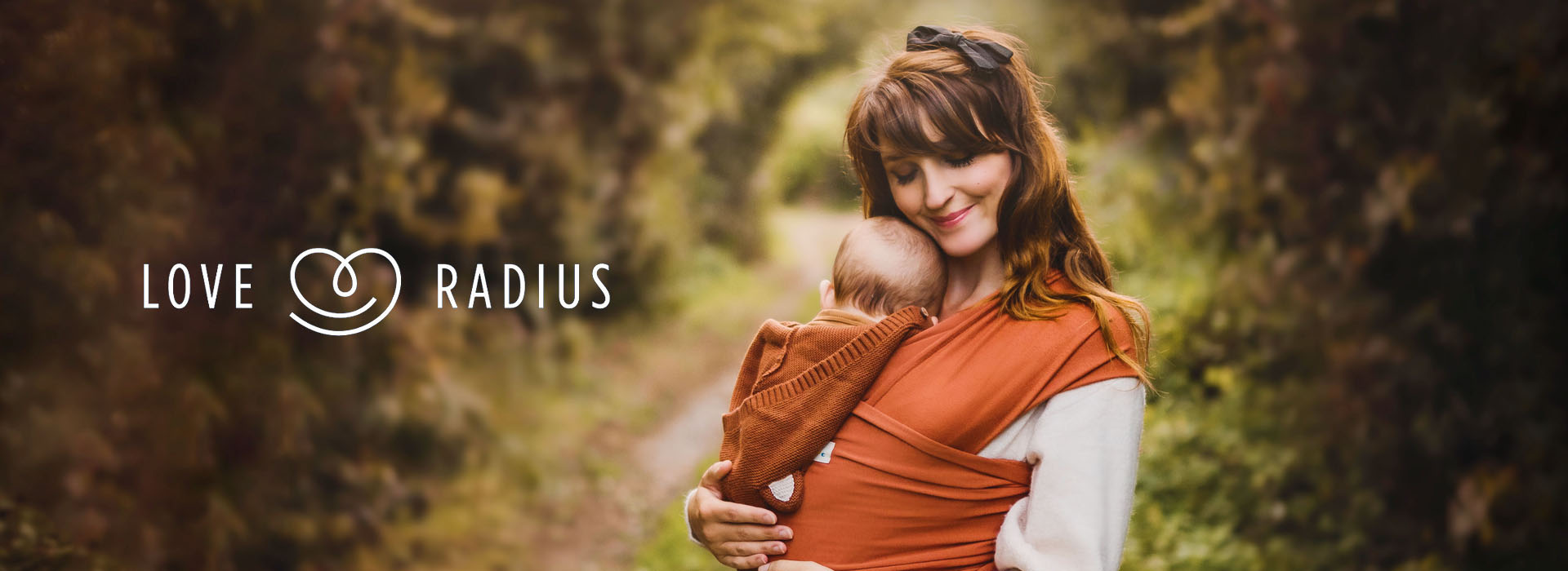 Nainen pitää vauvaa kantoliinassa metsäalueella, logo vasemmalla.