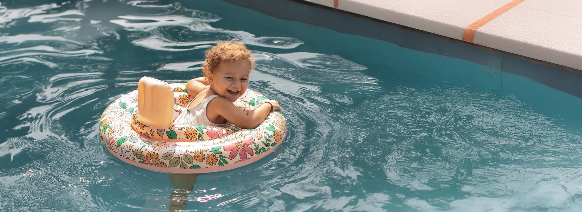 Hymyilevä lapsi kukkakuviollisessa uimarenkaassa, jossa on selkänoja. Kauden uutuudet.