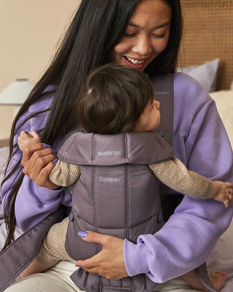 Nainen violetissa paidassa kantaa vauvaa kantorepussa sisällä.