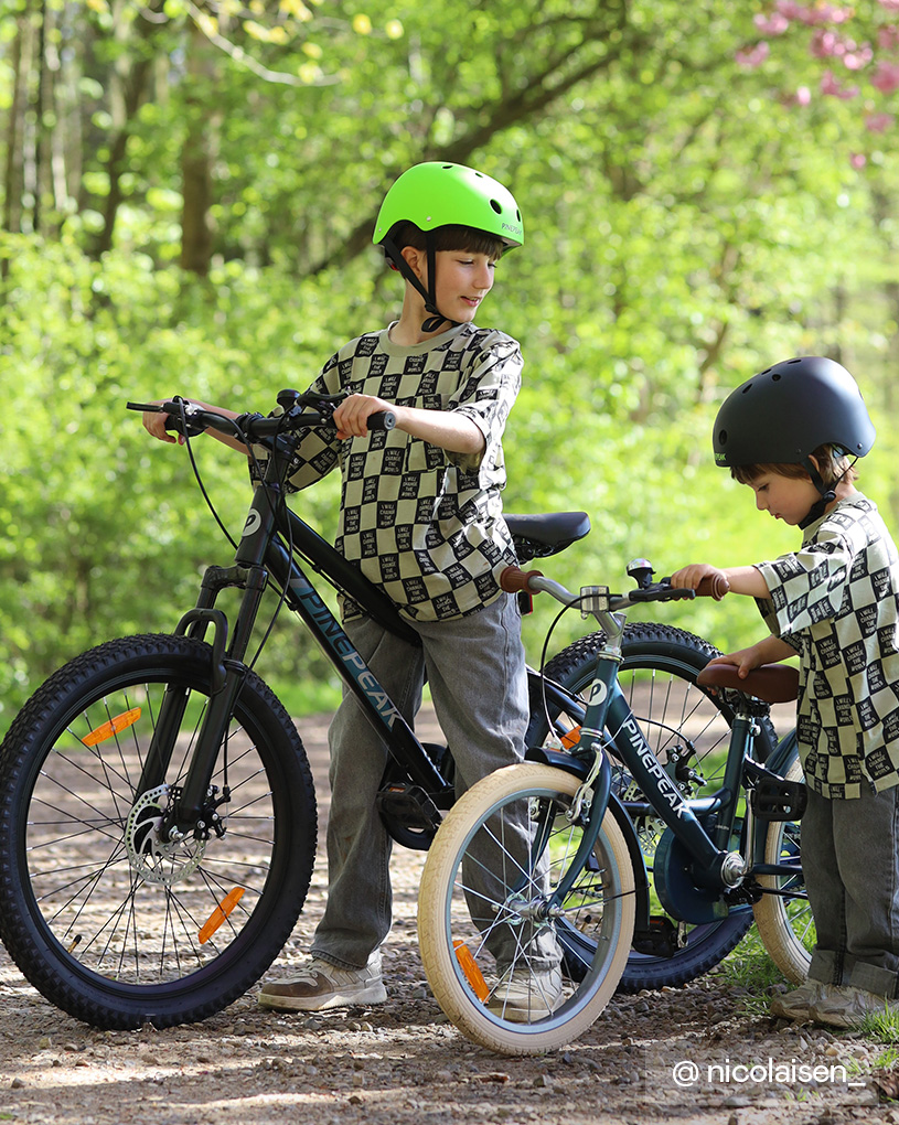 Kaksi kypäräpäistä lasta pyöräilevät vihreässä metsässä.