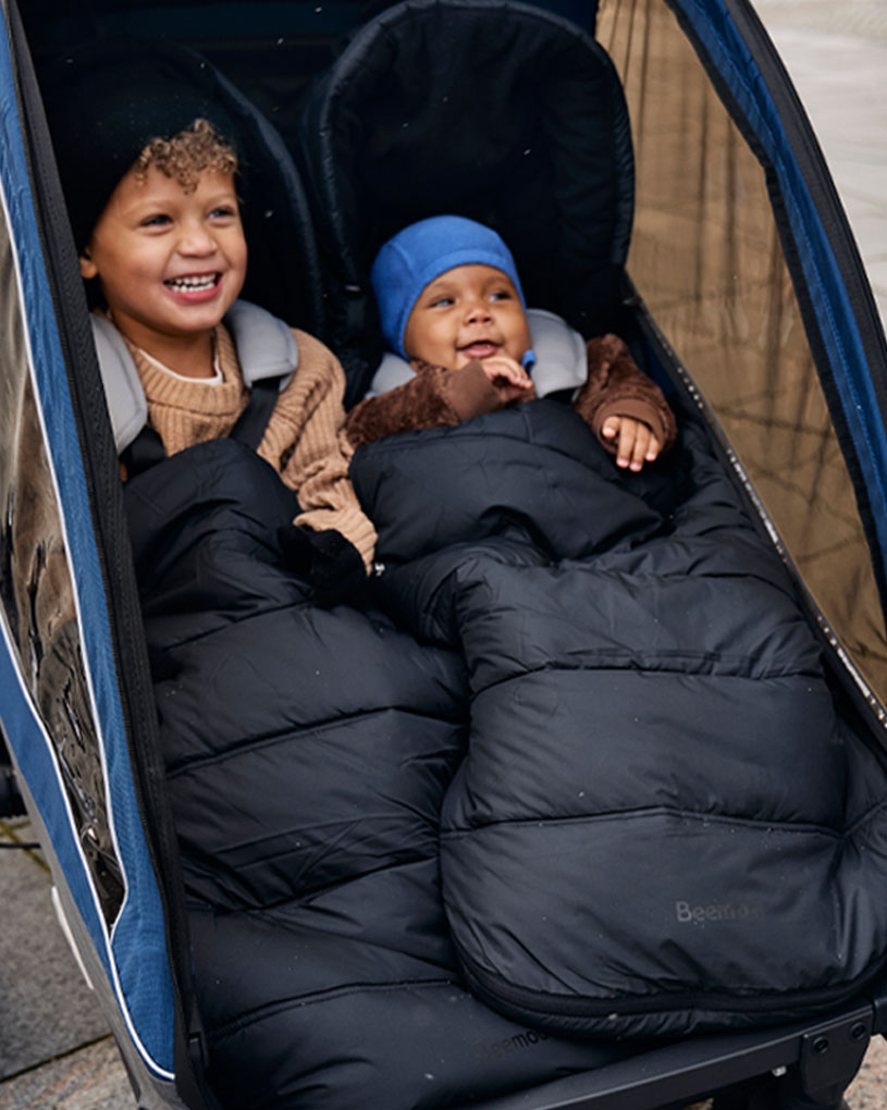 Kaksi iloista lasta makuupusseissa kaksosrattaissa.